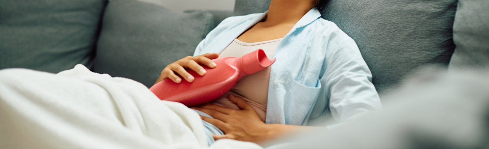 Eine Frau liegt auf dem Sofa und benutzt eine rote, medizinische Wärmflasche zur Wärmetherapie